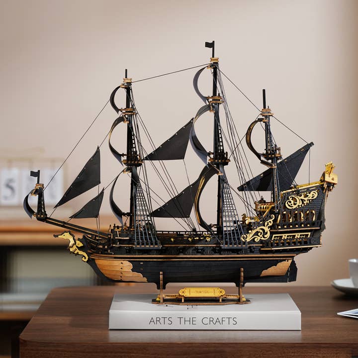 A detailed model of the DIY The Seahorse Barque Ship by Hands Craft, featuring black and gold colors with multiple masts, is displayed on a wooden table atop a stand labeled "ARTS THE CRAFTS" in soft lighting.