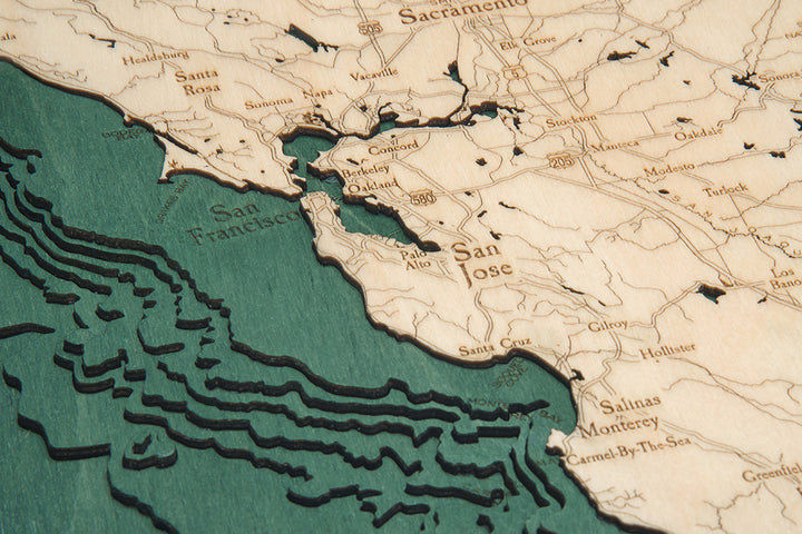 A close-up of the WoodChart California Coast 24.5" x 31" Black Frame map highlights the San Francisco Bay Area with detailed coastlines, labeled roads, and cities like San Francisco, San Jose, and Santa Rosa on a textured wooden surface.