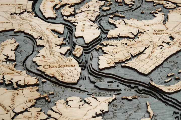 A close-up of the WoodChart CharlstonSC 24.5 x 31 Grey Frame showcases intricate laser-cut layers of Charleston, SC’s land, rivers, and harbor with labeled place names on a stunning wooden map.