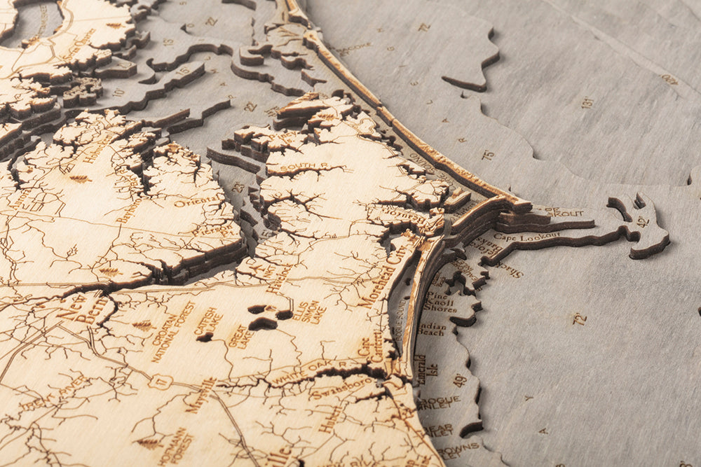 Close-up of the WoodChart North Carolina Coast 24.5" x 31" map in a grey frame, showing detailed laser-etched coastlines, rivers, and cities like Norfolk, Cape Henry, and Cape Lookout along the eastern United States.