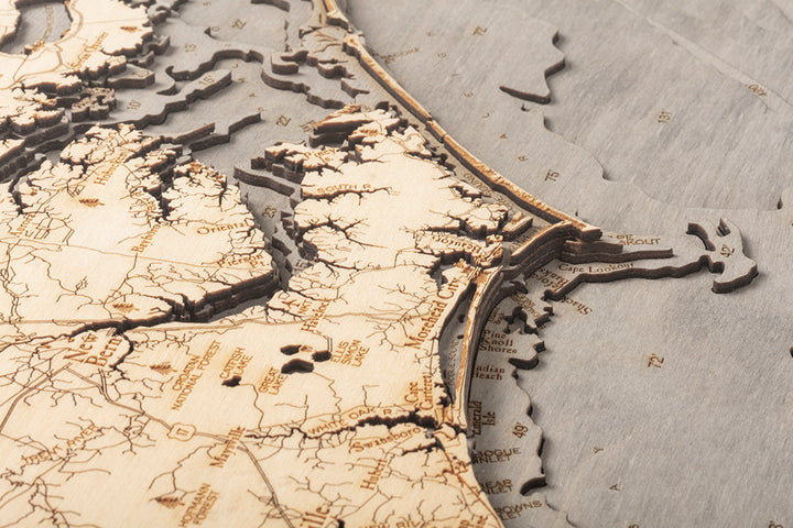 Close-up of the WoodChart North Carolina Coast 24.5" x 31" map in a grey frame, showing detailed laser-etched coastlines, rivers, and cities like Norfolk, Cape Henry, and Cape Lookout along the eastern United States.
