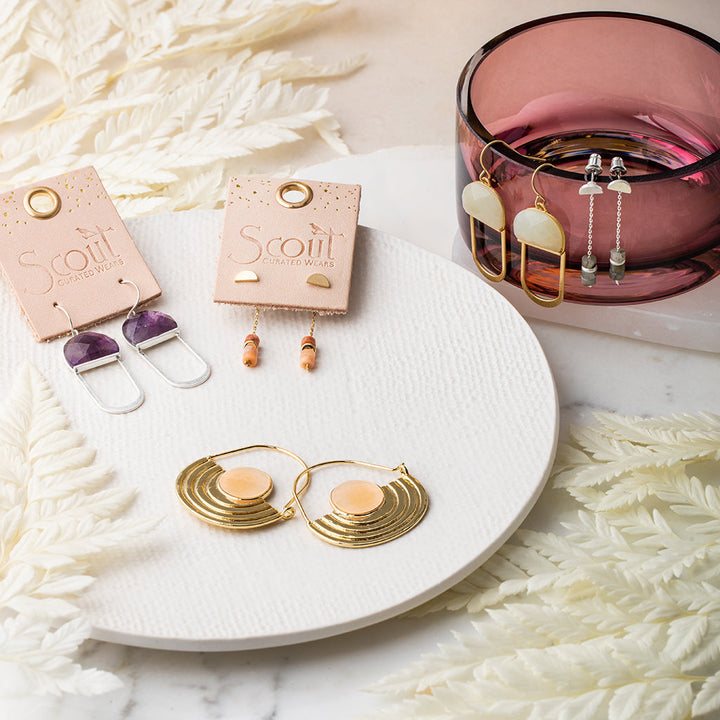 A white ceramic plate displays gold and pink circular earrings alongside dangling two-tone ones. On Scout-branded cards, hypoallergenic earrings rest gracefully. A pink glass holds more pairs, some with 14K gold dipped elegance. Fern leaves adorn the marble-like backdrop.