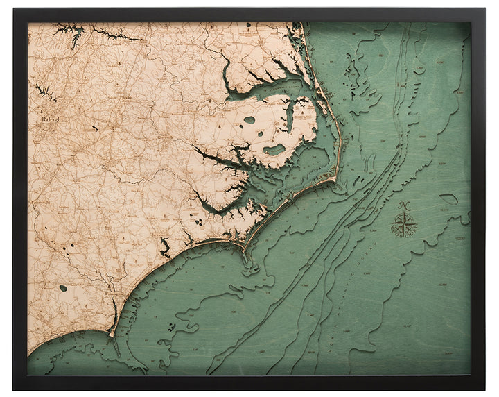 The WoodChart North Carolina Coast 24.5" x 31" Black Frame displays an intricately carved wooden map in natural wood and teal tones, featuring the region's shoreline, bodies of water, and inland details.