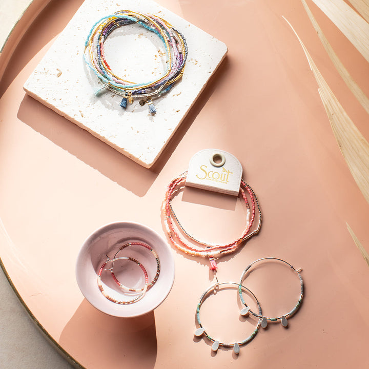 A collection of colorful beaded jewelry is arranged on a light pink tray. The items include stacking bracelets, a pair of hoop earrings with small dangling beads, another pair of hoop earrings with charm details, and the "Turquoise Multi Silver Miyuki Bracelet Trio" by Scout Jewelry, featuring Miyuki Delica beads on a branded tag.