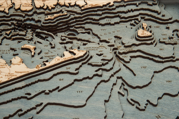 A close-up of the WoodChart Long Island Sound 13.5" x 31" map in a white frame, featuring layered wood, beige and blue tones, labeled locations, depth markings, and detailed land and underwater topography.