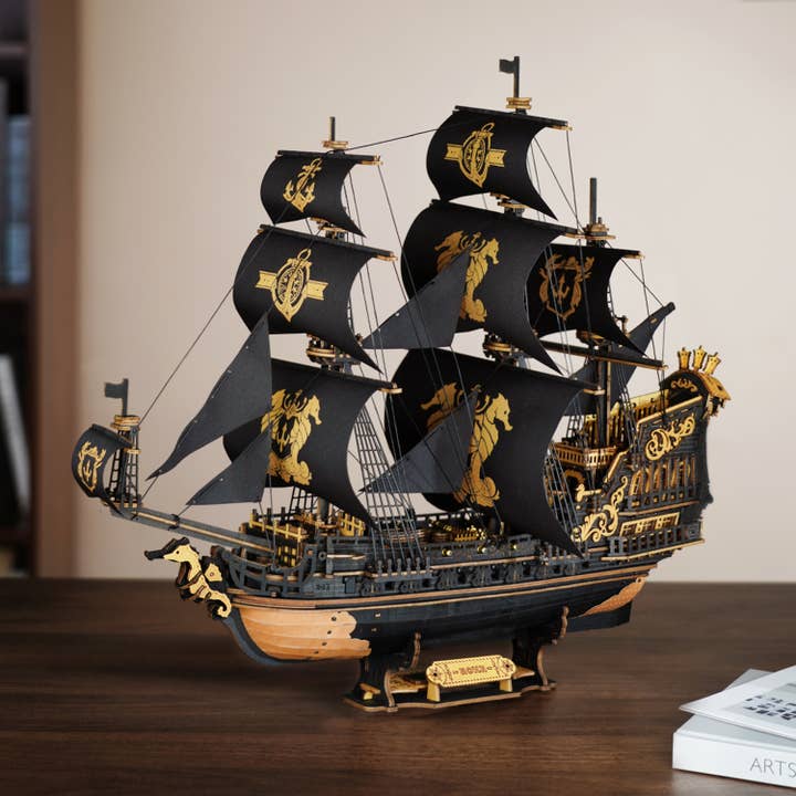The Hands Craft DIY The Seahorse Barque Ship, a black and gold pirate ship model with ornate decorations and multiple sails, sits on a wooden table beside a white book.
