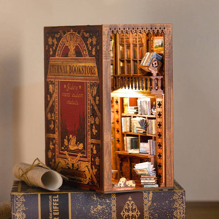 The Hands Craft DIY Book Nook Kit: Eternal Bookstore with Dust Cover opens to reveal a miniature, illuminated bookstore scene—shelves, books, intricate details—making it a unique literary gift.