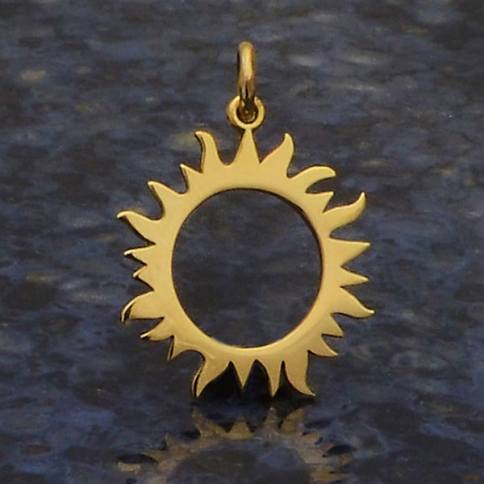 Nina Designs' Bronze Eclipse Sun Charm, featuring a gold sun shape with a hollow center and stylized pointed rays, elegantly rests on a textured dark surface, capturing the celestial mystery.