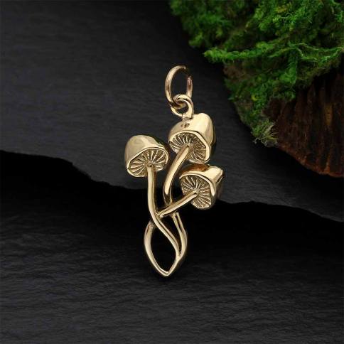 The Bronze Three Mushroom Charm by Nina Designs features a gold mushroom-shaped pendant with bronze tones and three intertwined stems against a dark background with green moss, making it an enchanting fairy tale gift or creative jewelry accessory.