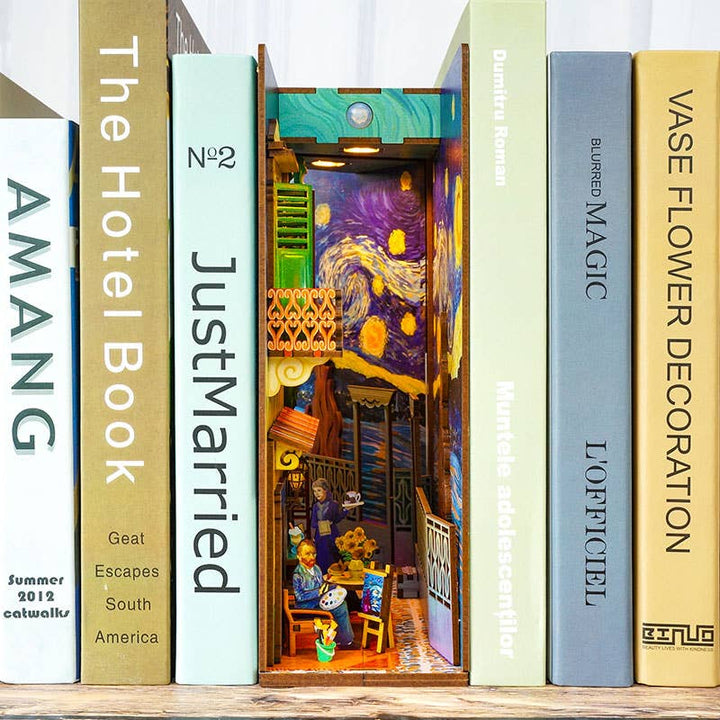A row of colorful books stands on a shelf, with Hands Craft’s "DIY Book Nook Kit: Vincent's World" featuring a detailed "Starry Night" diorama and mini figures—a creative décor piece nestled between your books.