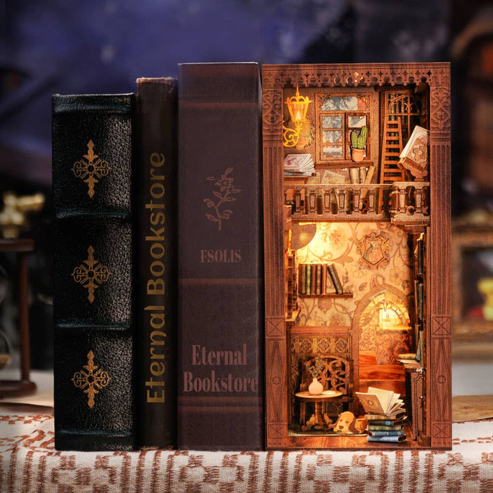 Three large book-shaped boxes sit side by side; one opens to reveal the Hands Craft DIY Book Nook Kit: Eternal Bookstore with Dust Cover—a warmly lit miniature bookstore scene with bookshelves, stairs, and cozy decor. The perfect literary gift for any book lover.