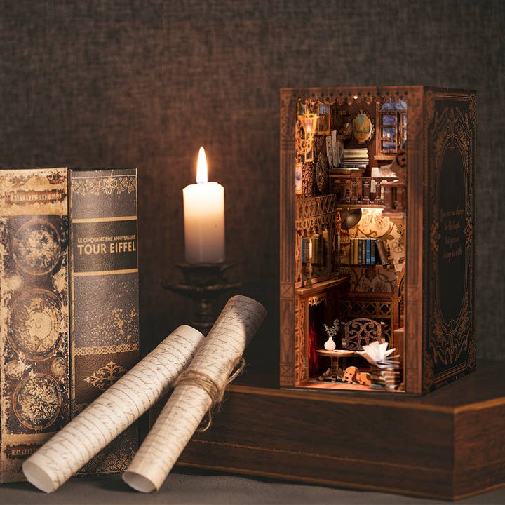 A candle, antique books, and scrolls sit beside the Hands Craft DIY Book Nook Kit: Eternal Bookstore with Dust Cover, featuring intricate miniature shelves, a desk, and warm lighting—perfect for creating a cozy library scene or unique literary gift.