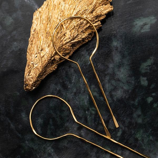 A pair of Equinox Keyhole Hoop Gold, hypoallergenic hairpins by Scout Jewelry, with circular ends lie on a dark green textured surface. Behind the nickel and cadmium-free hairpins is a gold, leaf-shaped decorative object.