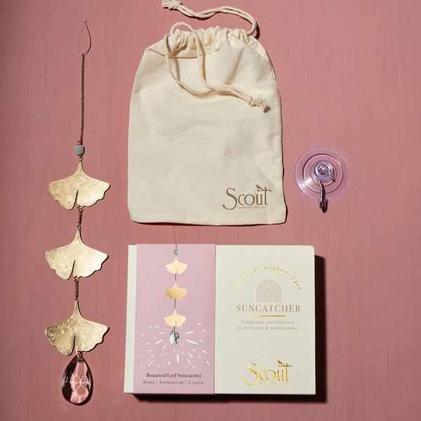 The Evil Eye Amethyst Suncatcher by Scout Jewelry features gold leaf accents and a sparkling pink crystal that reflects rainbows, displayed on a pink surface. It includes a matching box, beige drawstring pouch labeled "Scout," and a suction cup for hanging.