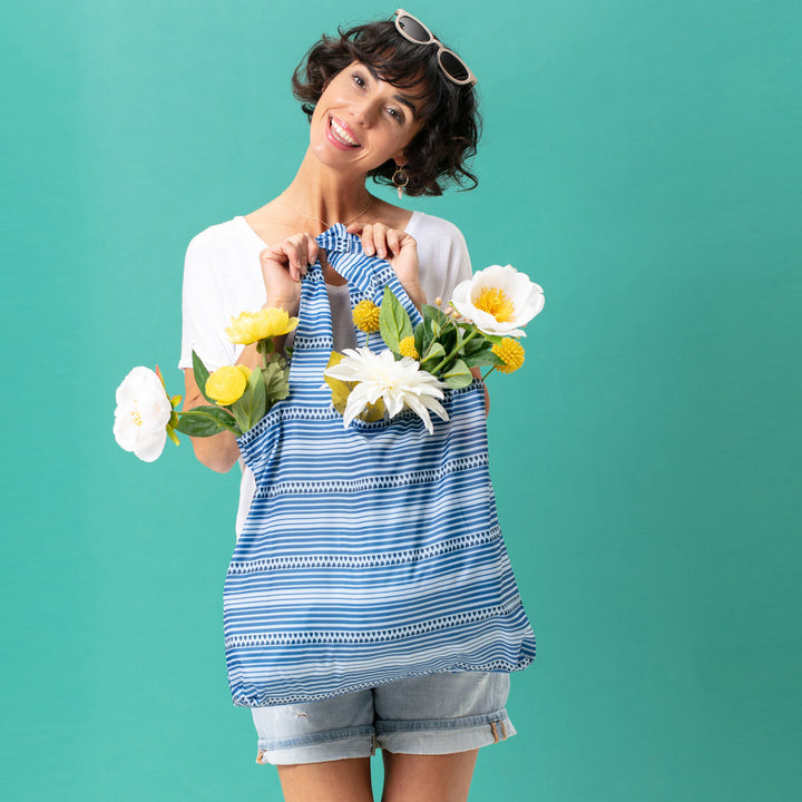 A woman in a white shirt and denim shorts smiles while holding a Bethany Reusable Bag from zzzRockFlowerPaper, filled with white and yellow flowers. She poses against a green background, sunglasses perched on her head, embodying an eco-friendly style.