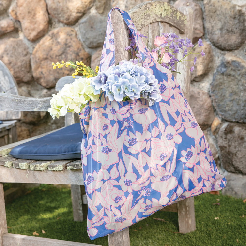 A Rosalie Reusable Bag in a floral pattern from zzzRockFlowerPaper, filled with pastel-colored flowers, hangs on an old wooden chair against a stone wall, creating a charming rustic outdoor setting. This reusable shopping bag not only enhances the scene but also supports an environmentally friendly lifestyle.