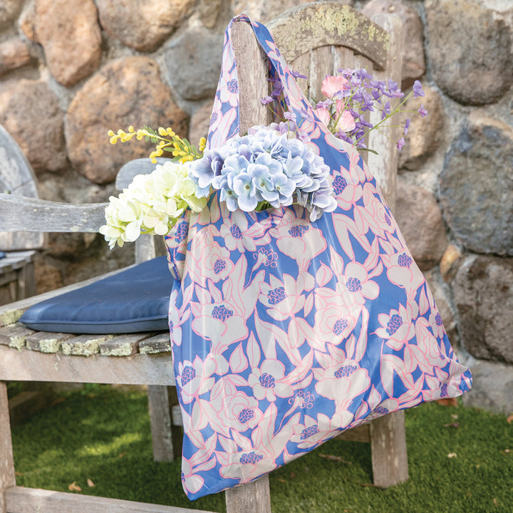 A Rosalie Reusable Bag in a floral pattern from zzzRockFlowerPaper, filled with pastel-colored flowers, hangs on an old wooden chair against a stone wall, creating a charming rustic outdoor setting. This reusable shopping bag not only enhances the scene but also supports an environmentally friendly lifestyle.