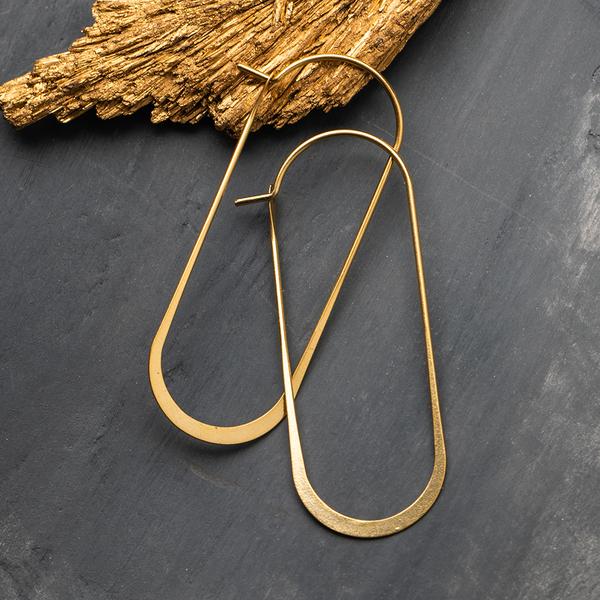 The Cosmic Oval Gold earrings by Scout Jewelry, crafted in 18K Gold Vermeil with a teardrop shape, are elegantly displayed on a dark textured surface with a piece of gold leaf in the background. These hypoallergenic earrings are both nickel and cadmium free, offering comfortable and stylish wear for everyday use.