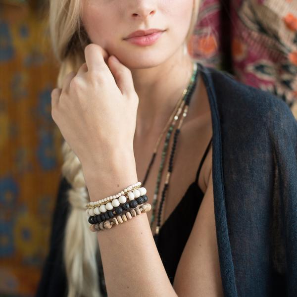A person with long, blonde hair styled in a braid is shown from shoulders up. They are dressed in a dark, open-front garment over a black top and wear multiple gemstone bracelets, including an Amazonite Stone Stacking Bracelet by Scout Jewelry, and layered necklaces featuring semi-precious gemstone beads. The background displays colorful fabric patterns.
