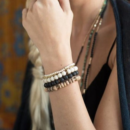 A person with light skin, wearing a sleeveless black top and a black shawl, showcases an arm adorned with several Scout Jewelry's Lava Stone Stacking Bracelets in shades of white, black, and beige. These gemstone bracelets add to their unique look. The scene is indoors, against a blurred colorful background.