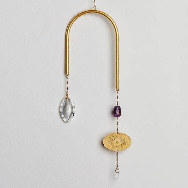 The Evil Eye Amethyst Suncatcher by Scout Jewelry features a minimalist gold metallic arch, silver leaf, radiant purple gem, and gold pendant with sun and eye designs. Its crystals reflect rainbows elegantly against the plain white background.