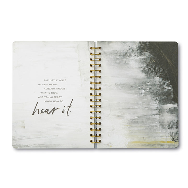A "You are here to do incredible things" spiral notebook by Compendium displays abstract white and dark gray brushstrokes on the right-hand page, while the left-hand page features an inspiring message in simple handwriting: "The little voice in your heart already knows what's true and you already know how to hear it.