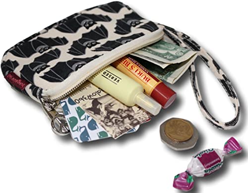 The Bungalow 360 Tiny Wristlet Bat, a small zippered pouch with a unique black and white pattern, sits open, revealing various items including cash, a tube of lotion, chapstick, and a small notebook. Coins and a wrapped candy are placed next to the charming cotton canvas wristlet.