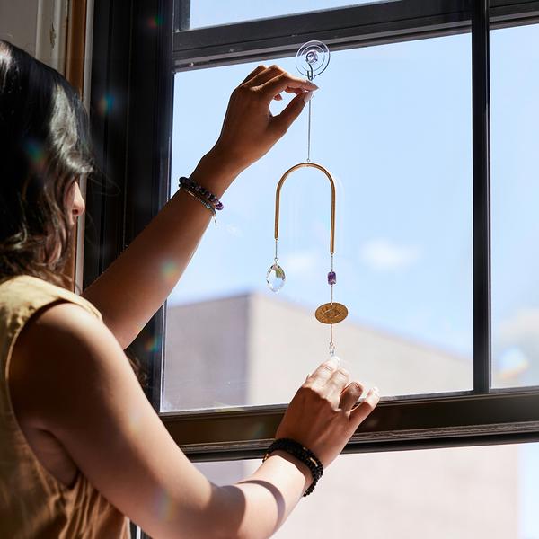 A person with long hair and bracelets hangs the Scout Jewelry Evil Eye Amethyst Suncatcher on a window using a suction cup, its crystal-adorned glass casting subtle rainbows. A building and blue sky are visible outside.