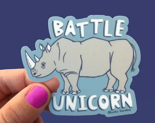 A hand with purple nail polish holds a Rhino Battle Unicorn Sticker from Lucky Sardine, featuring an illustration of a rhino with the text "Battle Unicorn" on a blue background. This durable vinyl sticker is weatherproof, ensuring your "Battle Unicorn" stands strong through any conditions.