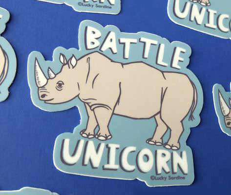 Introducing the Rhinoceros Rhino Battle Unicorn Sticker by Lucky Sardine! This weatherproof sticker showcases an illustration of a rhinoceros standing on a light blue background with the words "BATTLE UNICORN" written above. Made of durable vinyl, this playful design features a charming white border.