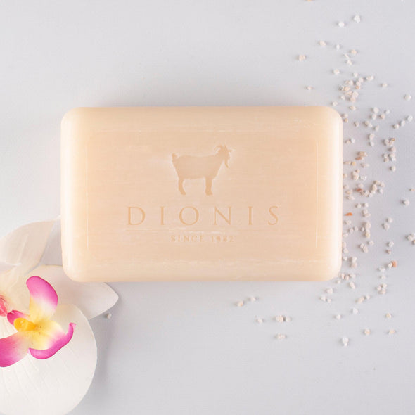 A bar of Sea Treasures Soap by Dionis, weighing 6oz and colored beige with shea butter, is embossed with a goat emblem and the text "DIONIS SINCE 1982," rests gracefully on a light surface. Around it, scattered pink and white flower petals combine with small salt granules to create an inviting, spa-like ambiance.