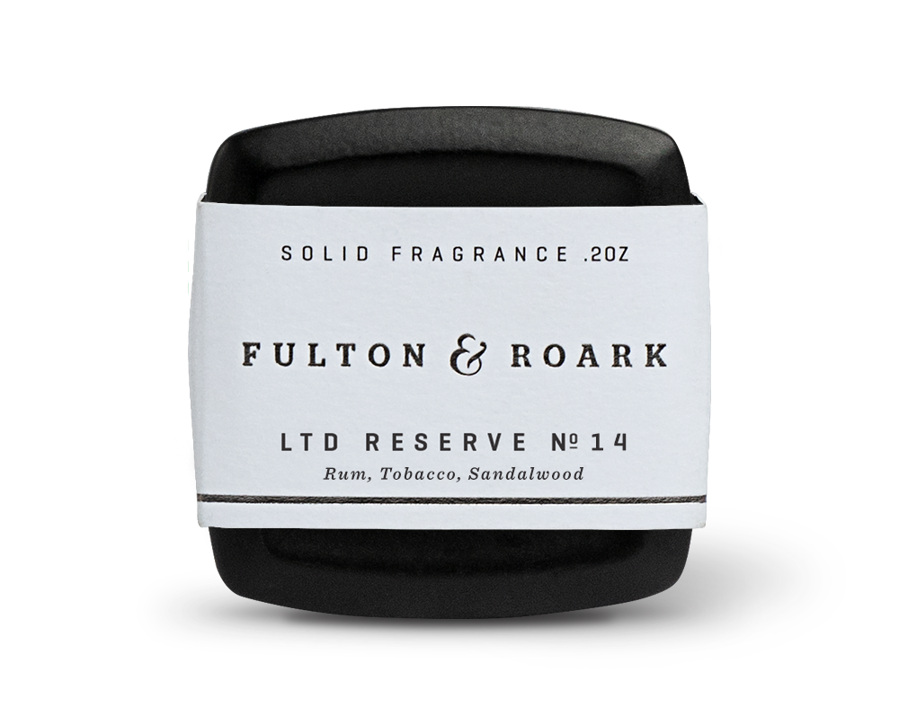 A small, sleek black container of solid fragrance from Fulton & Roark. The label reads "Calle Ocho .2oz," and the scent details below conjure up notes of Rum Tobacco, Sandalwood. The container lies on a white surface, casting a slight shadow reminiscent of Calle Ocho's vibrant charm.