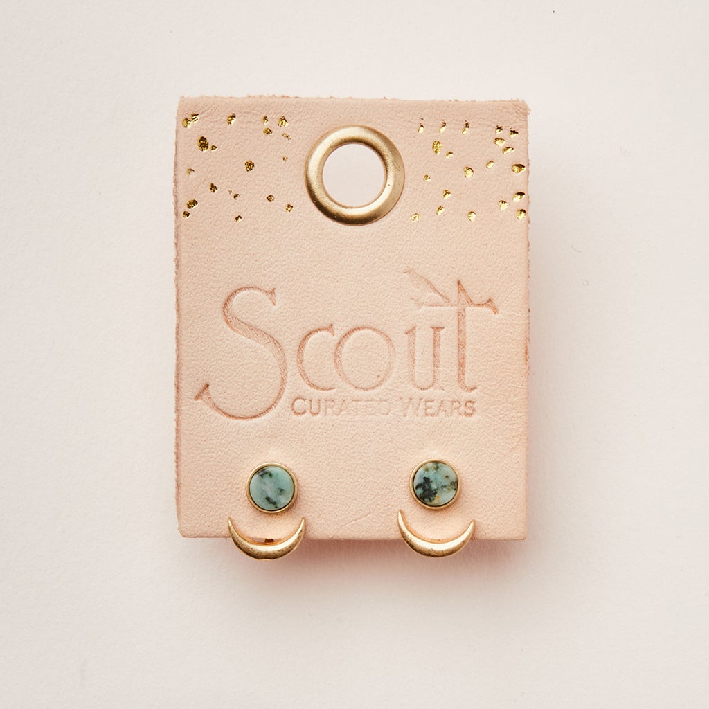 A pink card with "Scout Jewelry" embossed on it holds a pair of hypoallergenic stud earrings. The earrings, named Moon Phase African Turq/Gold, feature adjustable brass crescent shapes with small turquoise stones above them. The card also has a round hole at the top and gold speckles.