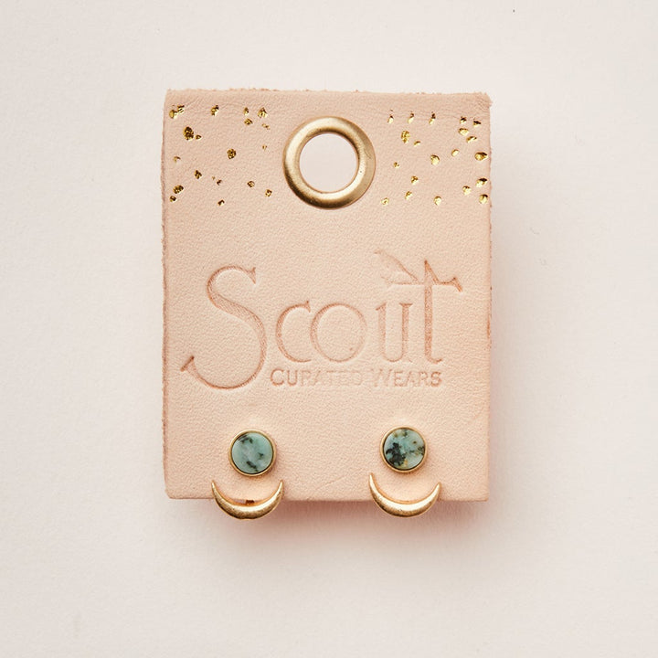 A pink card with "Scout Jewelry" embossed on it holds a pair of hypoallergenic stud earrings. The earrings, named Moon Phase African Turq/Gold, feature adjustable brass crescent shapes with small turquoise stones above them. The card also has a round hole at the top and gold speckles.