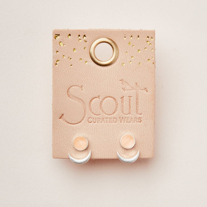 A pair of Moon Phase Sunstone/Silver earrings is showcased on a beige leather card adorned with gold specks and a small metal ring at the top. The card is embossed with the words "Scout Jewelry." These hypoallergenic earrings boast a minimalist design featuring round peach-colored studs and adjustable brass crescents.