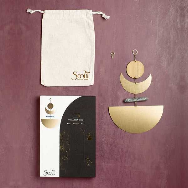 The Moonrise Labradorite Wall Hanging by Scout Jewelry is showcased against a purple backdrop, highlighting geometric sculptural wall art. It comes with a beige pouch featuring the "Scout" branding, a black and white box adorned with golden accents, and includes metallic pieces in natural brass for an elegant finish.