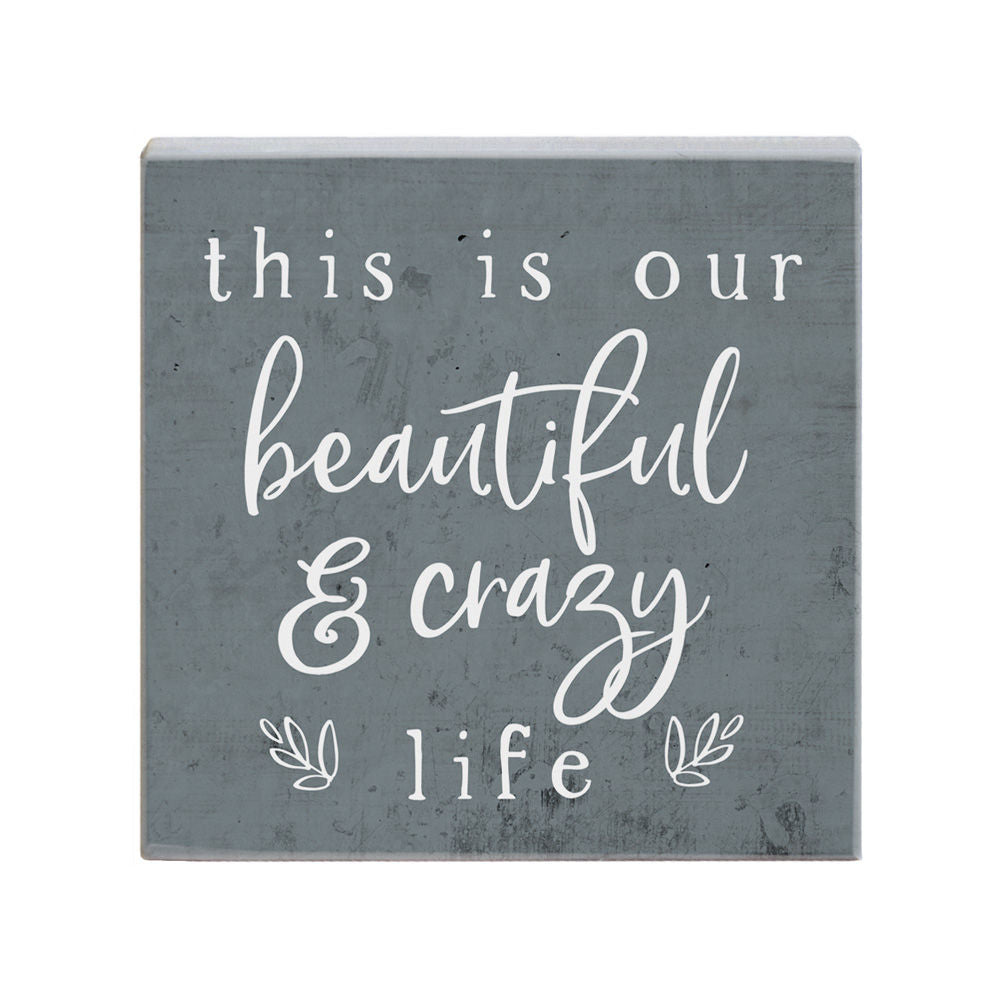 The "Crazy Life" plaque by SincereSurroundings is a handcrafted square piece featuring an inspirational quote, "this is our beautiful & crazy life," elegantly displayed in white cursive and print on a gray background. Its small decorative elements enhance the lower corners, making it a charming addition to any room and ideal for shelf display.