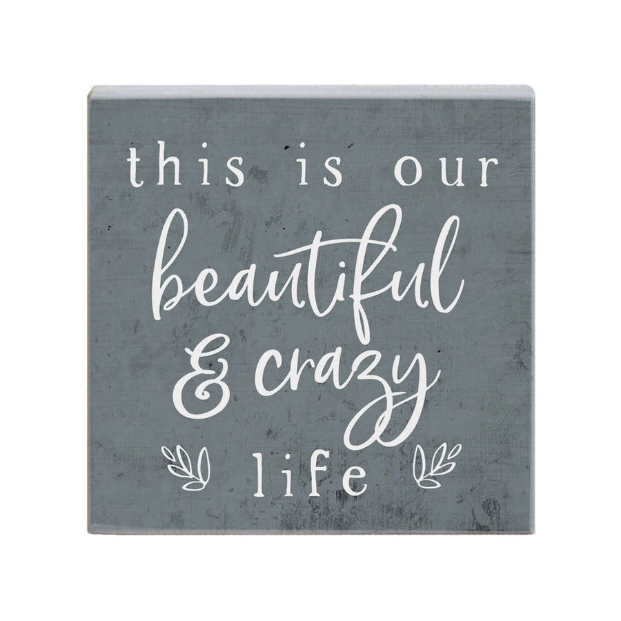 The "Crazy Life" plaque by SincereSurroundings is a handcrafted square piece featuring an inspirational quote, "this is our beautiful & crazy life," elegantly displayed in white cursive and print on a gray background. Its small decorative elements enhance the lower corners, making it a charming addition to any room and ideal for shelf display.