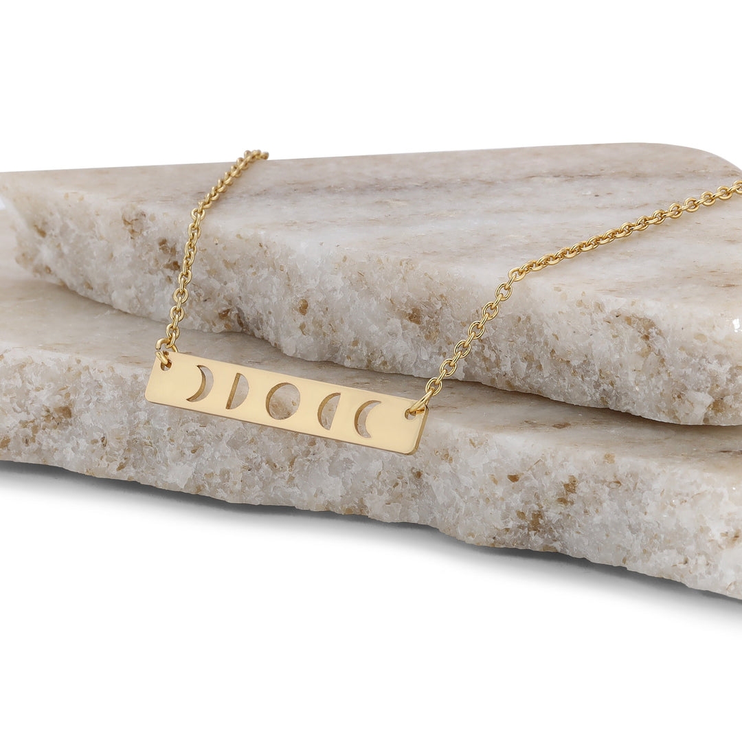 The Moon Phase Necklace Gold by LuckyFeather is a stunning piece for astrology lovers, crafted from 14k gold-dipped brass with a rectangular pendant featuring moon phase cutouts, elegantly presented on stacked marble slabs.