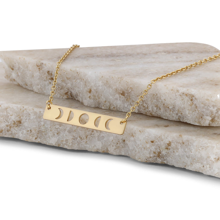 The Moon Phase Necklace Gold by LuckyFeather is a stunning piece for astrology lovers, crafted from 14k gold-dipped brass with a rectangular pendant featuring moon phase cutouts, elegantly presented on stacked marble slabs.