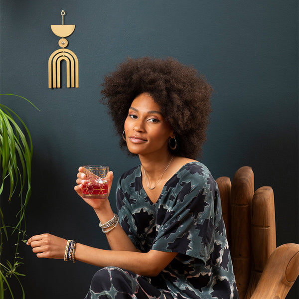 A woman with curly hair sits in a chair holding a drink. She is adorned in a patterned outfit, bracelets, and hoop earrings. Behind her, the Moonrise Labradorite Wall Hanging by Scout Jewelry adds a touch of modern geometric elegance to the dark background, and a plant is partially visible on the side.