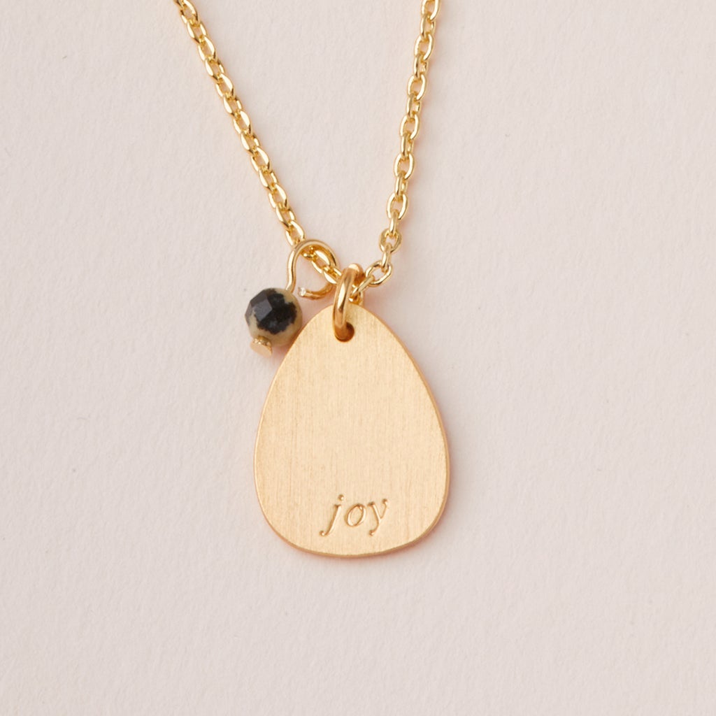 The Intention Neck Labradorite/Gold necklace by Scout Jewelry features a teardrop-shaped pendant engraved with "joy," a small black bead, and hangs from an adjustable brass chain, suitable for any occasion.