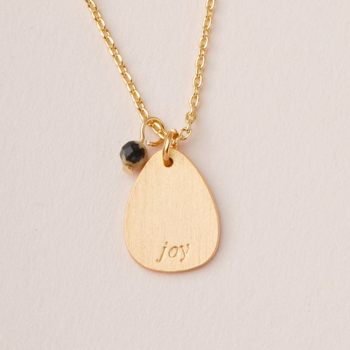 The Intention Neck Labradorite/Gold necklace by Scout Jewelry features a teardrop-shaped pendant engraved with "joy," a small black bead, and hangs from an adjustable brass chain, suitable for any occasion.