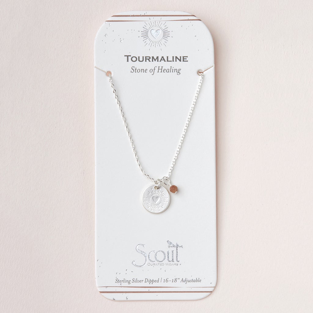 The "Intention Neck Tourmaline/Silver" by Scout Jewelry is a silver necklace with a round pendant and small stone, packaged on a "Tourmaline Stone of Healing" card. It features semi-precious gemstones, an engraved charm, and an adjustable 16-18 inch sterling silver dipped chain.