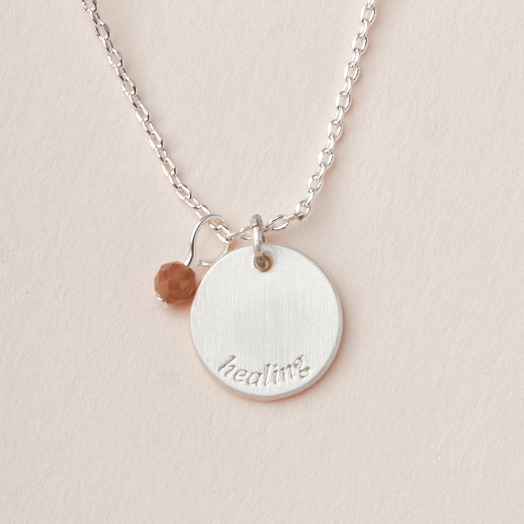 Scout Jewelry's "Intention Neck Tourmaline/Silver" features a silver necklace with an adjustable chain, a round pendant engraved with "healing" charms, and a small brown bead, elegantly displayed on a light beige surface.