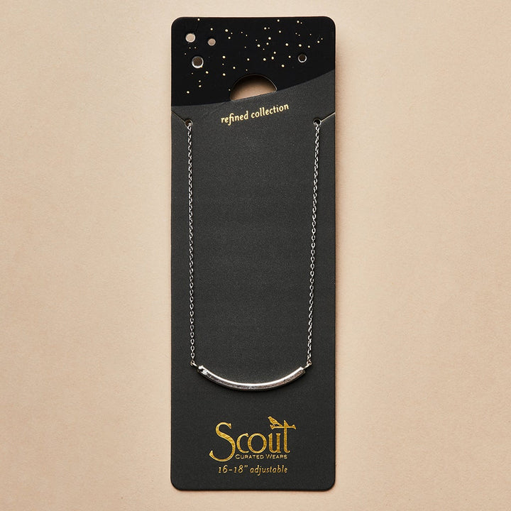 The "Refined Neck Comet/Silver" by Scout Jewelry is a silver necklace featuring a curved bar design with CZ stones. It comes in black and gold packaging with a starry pattern, labeled "Scout Curated Wears," set against a soft beige background displaying the text "refined collection.