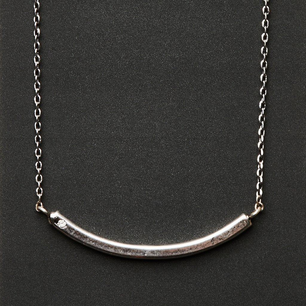 The Refined Neck Comet/Silver by Scout Jewelry is a minimalist piece with a curved bar-shaped pendant crafted from 925 silver. Its delicate chain enhances the sleek, polished look set against a dark background.