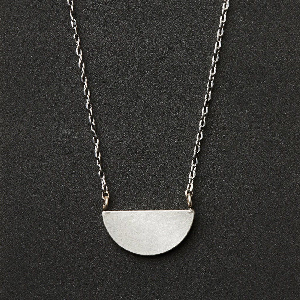 The Refined Neck Half Moon/Silver by Scout Jewelry features a simple adjustable silver chain with a flat, matte half-circle pendant at the center, displayed against a black background.