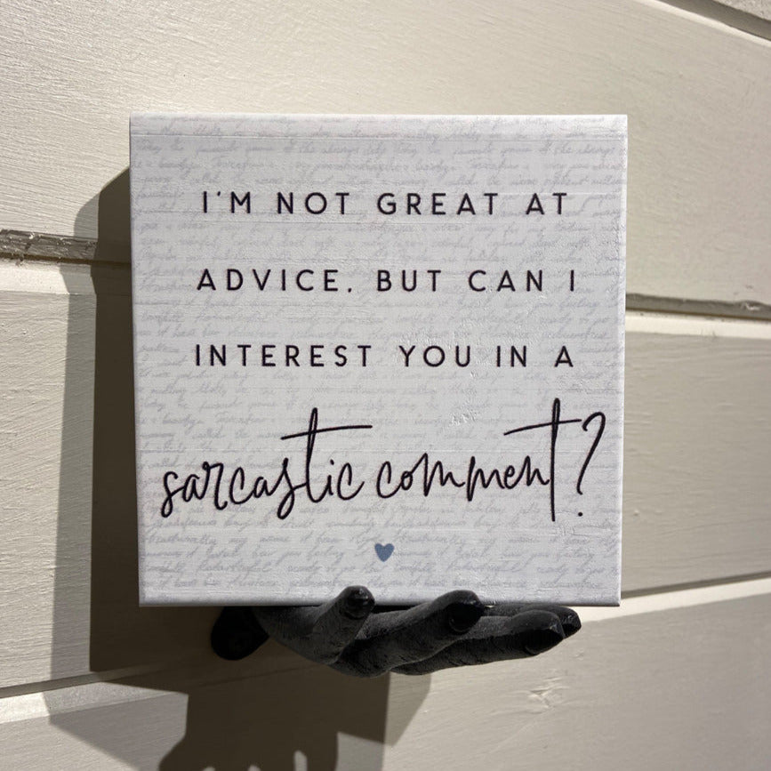 Close-up of a handcrafted sign from SincereSurroundings, held by a decorative black hand against a light wooden wall. The "Sarcastic Comment" sign features the phrase, "I'm not great at advice, but can I interest you in a sarcastic comment?" in black script and print fonts with a small heart design at the bottom. It's perfect for your shelf display.