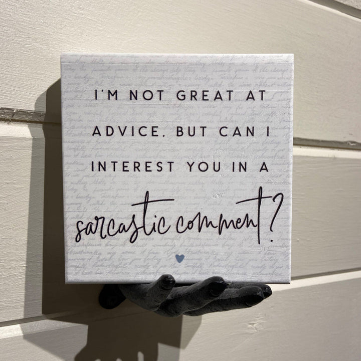 Close-up of a handcrafted sign from SincereSurroundings, held by a decorative black hand against a light wooden wall. The "Sarcastic Comment" sign features the phrase, "I'm not great at advice, but can I interest you in a sarcastic comment?" in black script and print fonts with a small heart design at the bottom. It's perfect for your shelf display.